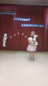 Dziewczynka stoi na scenie i śpiewa piosenkę do mikrofonu.