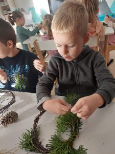 Przedszkolaki siedzą przy stoliku i wykonują wianki z wierzby płaczącej, szyszek, trawy ozdobnej i innych ekologicznych materiałów.