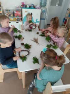 Przedszkolaki siedzą przy stoliku i wykonują wianki z wierzby płaczącej, szyszek, trawy ozdobnej i innych ekologicznych materiałów.