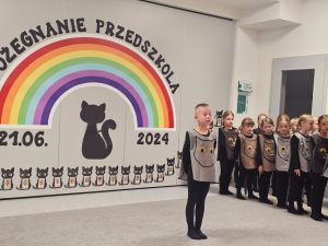 Dzieci ubrane na czarno w kocich kamizelkach przedstawiają program słowno-taneczny na tle tęczy.