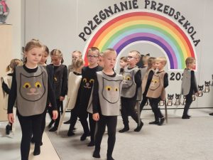Dzieci ubrane na czarno w kocich kamizelkach przedstawiają program słowno-taneczny na tle tęczy.