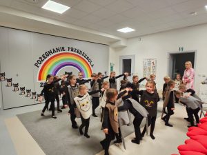 Dzieci ubrane na czarno w kocich kamizelkach przedstawiają program słowno-taneczny na tle tęczy.