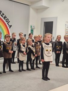 Dzieci ubrane na czarno w kocich kamizelkach przedstawiają program słowno-taneczny na tle tęczy.