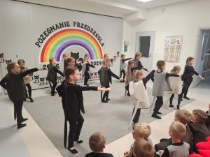 Dzieci ubrane na czarno w kocich kamizelkach przedstawiają program słowno-taneczny na tle tęczy.