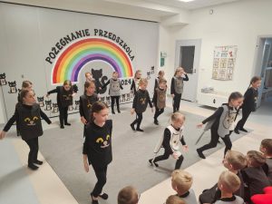 Dzieci ubrane na czarno w kocich kamizelkach przedstawiają program słowno-taneczny na tle tęczy.