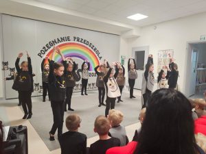 Dzieci ubrane na czarno w kocich kamizelkach przedstawiają program słowno-taneczny na tle tęczy.