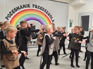 Dzieci ubrane na czarno w kocich kamizelkach przedstawiają program słowno-taneczny na tle tęczy.