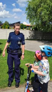 Dzieci na rowerach i kaskach w Miasteczku Ruchu Drogowego słuchają poleceń pana Policjanta i pani Policjant, jak prawidłowo poruszać się po ulicy.