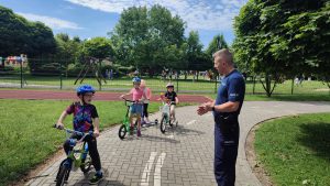 Dzieci na rowerach i kaskach w Miasteczku Ruchu Drogowego słuchają poleceń pana Policjanta i pani Policjant, jak prawidłowo poruszać się po ulicy.