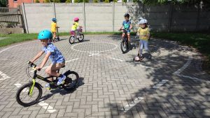 Dzieci na rowerach i hulajnogach kreślą ósemki na drodze. Doskonalą jazdę na rowerze i hulajnodze.