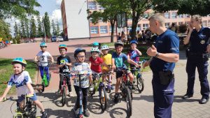 Dzieci na rowerach i kaskach w Miasteczku Ruchu Drogowego słuchają poleceń pana Policjanta i pani Policjant, jak prawidłowo poruszać się po ulicy.