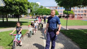Dzieci jadą na rowerach i hulajnogach po ulicach Miasteczka Ruchu Drogowego. Asystuje im policjant.
