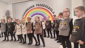Dzieci ubrane na czarno w kocich kamizelkach przedstawiają program słowno-taneczny na tle tęczy.