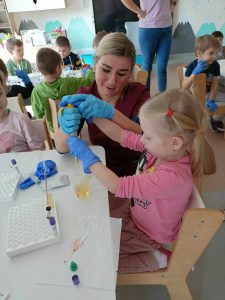 Pani Sylwia pomaga dziewczynce wykonać zadanie - wlanie soku do próbówki. Dziewczynka siedzi przy stoliku. Obydwie mają na rękach rękawiczki ochronne.