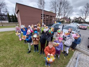 Dzieci trzymają koszyczki wielkanocne w rękach, stoją przed budynkiem przedszkola i uśmiechają się. W koszyczkach są słodycze.