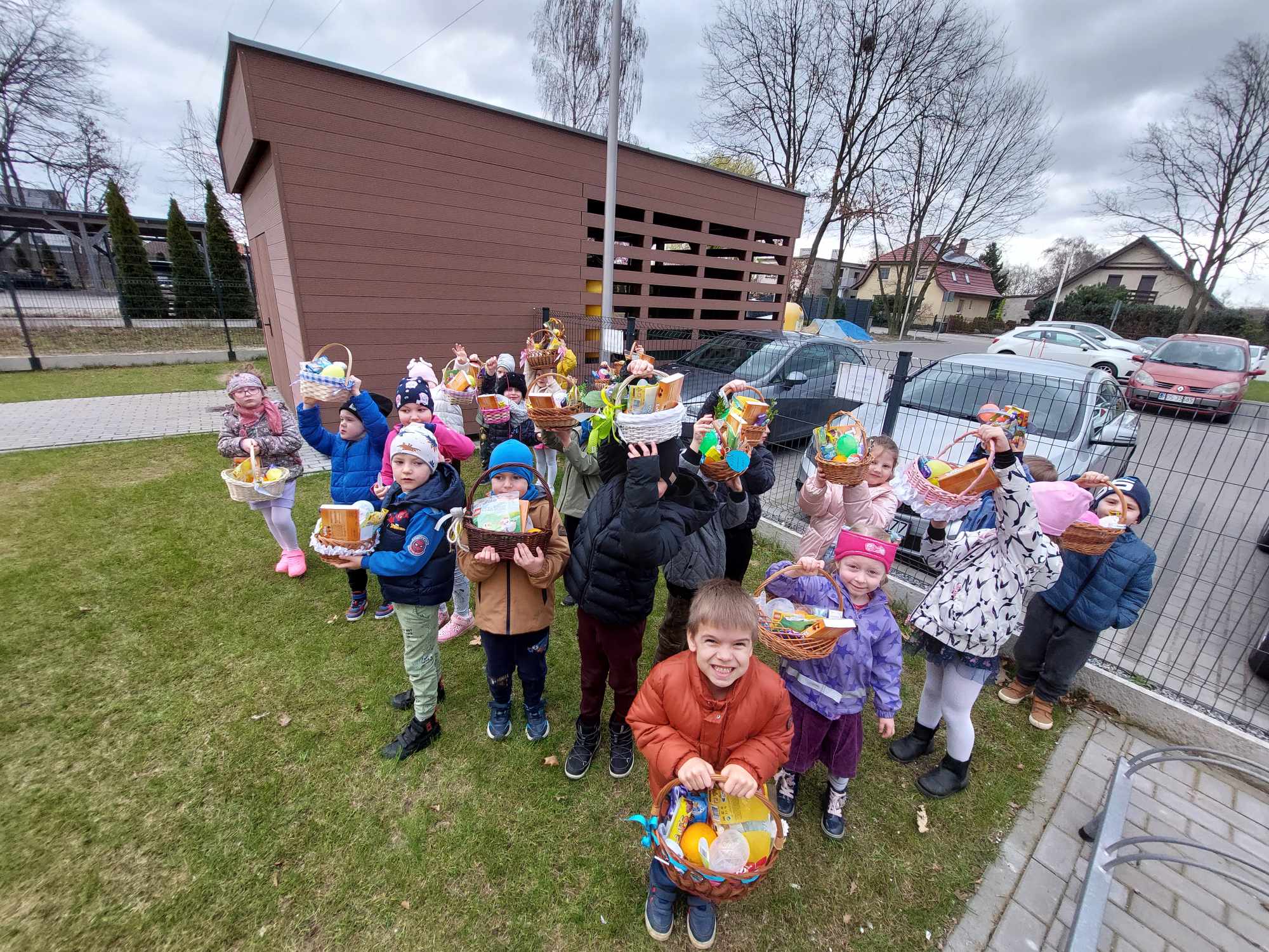 Dzieci trzymają koszyczki wielkanocne w rękach, stoją przed budynkiem przedszkola i uśmiechają się. W koszyczkach są słodycze.