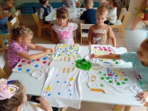 jeden chłopiec i cztery dziewczynki siedza przy stoliku i robia farbkami na koszulce kolorowe kropki
