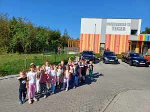Grupa dzieci stojących przed budynkiem przedszkola machających rękami.