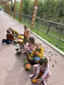 Dzieci siedzą obok ścieżki sensorycznej i rękoma dotykają jesienne dary- kasztany, dynie, patyki, szyszki i liście.