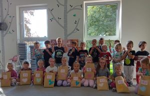 Grupa dzieci ustawiona w dwóch rzędach na tle okien. Dziewczynki siedzą na dywanie przed sob maja upominki w papierowych ozdobionych torbach. Za nimi stoja chłopcy i trzymaja w ręce wafelka.