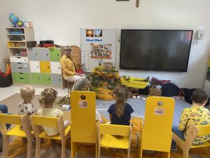 Dzieci i Pani siedzą na krześle i patrzą na tablicę. m