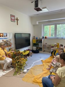 Dzieci i Pani siedzą na krześle i pokazują ruchy do piosenki.