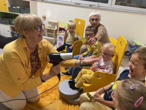 Dzieci siedzą na krześle. Pani kieruje szuszarkę i stronę dzieci i rozrzuca liście.