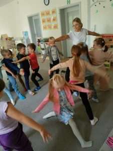 ZAJĘCIA Z JOGI. DZIECI WRAZ Z PANIĄ STOJĄ NA DYWANIE W SALI PRZEDSZKOLNEJ I MAJĄ ROZŁOŻONE NA BOKI RĘCE. ĆWICZĄ WYBRANE POZYCJE Z JOGI.