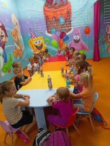 DZIEŃ PRZEDSZKOLAKA 2024. GRUPA DZIECI SIEDZI PRZY JEDNYM STOLE I KAŻDE MA PRZED SOBĄ BUTELKĘ Z NAPOJEM. W TLE KOLOROWA ŚCIANA Z POSTACIAMI Z BAJEK SPONGEBOB I DUŻY TORT - RĘCZNIE MALOWANE.