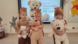 Troje dzieci ubranych na brązowo trzymają w rękach pluszowego misia i pozują do wspólnego zdjęcia na tle ekranu multimedialnego.