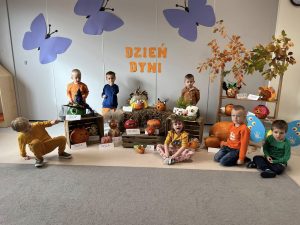 Dzieci stoją i pozują do zdjęcia na tle dekoracji pośród dyń i napisu Dzień Dyni. Prawie każde z nich ma element garderoby w kolorze pomarańczowym.