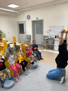Dzieci siedzą na krzesłach i podnoszą ręce do góry trzymając kolorową szarfę.