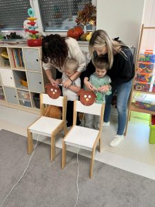 Dzieci stoją za krzesełkiem i z pomocą nauczyciela zwijają na patyczek nitkę, która przechodzi przez buźkę misia.
