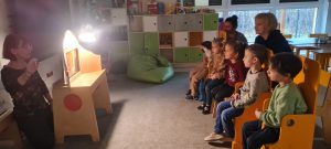 Grupa dzieci siedzi i patrzy na prezentowany przez Panią teatrzyk Kamishibai.