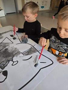 Dwoje dzieci ubranych na czarno maluje szarą farbą kontur dużego kota leżącego na białym stole.