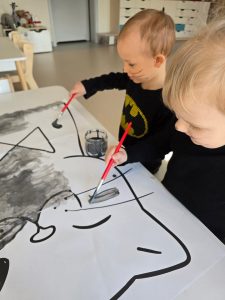 Dwoje dzieci ubranych na czarno maluje szarą farbą kontur dużego kota leżącego na białym stole.