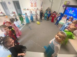 Dzieci w strojach karnawałowych stoi w dużym kole na szarym dywanie.