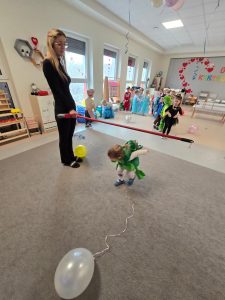 Dziewczynka w stroju karnawałowym przechodzi pod patykiem umieszczonym nad szarym dywanie. Patyk trzyma kobieta ubrana na czarno.