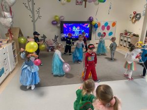 Duża grupa dzieci w kolorowych strojach tańczą na szarym dywanie, w rękach mają kolorowe balony.