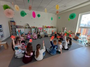 Dzieci siedzą w kole na dywanie trzymając w ręce czerwone kartki - tym sposobem odpowiadają na zadane przez nauczycielkę pytanie.