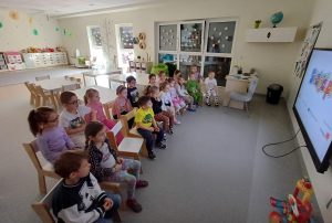 Przedszkolaki siedzą na krzesełkach przed ekranem monitora, oglądają film edukacyjny o bezpieczeństwie w sieci.