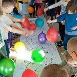 Dzieci stoją dookoła długiego arkusza białego papieru. W rączkach trzymają balony na sznurkach , którymi stemplują kolorową farbkę, rozmieszczoną na arkuszu.