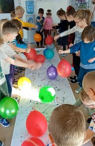 Dzieci stoją dookoła długiego arkusza białego papieru. W rączkach trzymają balony na sznurkach , którymi stemplują kolorową farbkę, rozmieszczoną na arkuszu.