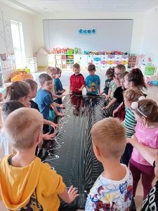 Grupa dzieci dookoła stolika , na którym jest położona czarna folia, a na niej biała farba, którą dzieci rozprowadzają rączkami