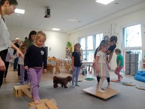 Dzieci przechodzą prze różne akcesoria ustawione na dywanie, w formie toru przeszkód i placu zabaw.