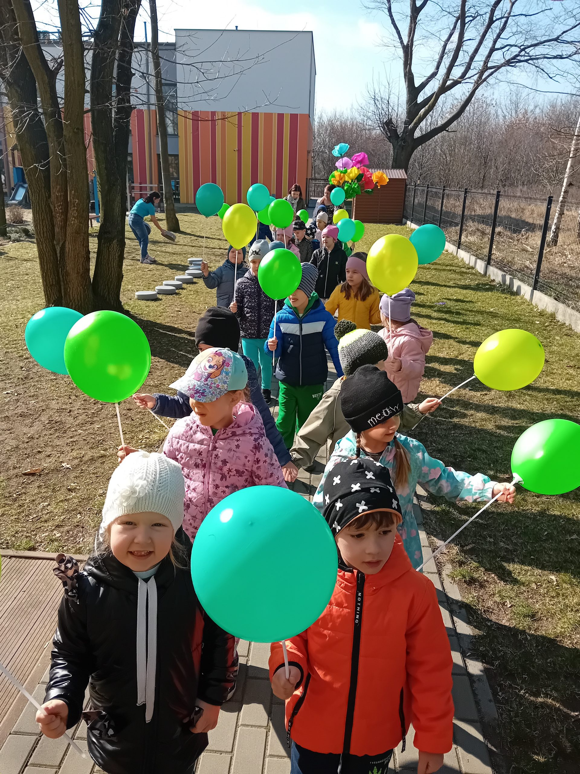 Grupa dzieci spaceruje w parach z zielonymi balonikami po przedszkolnym placu zabaw.