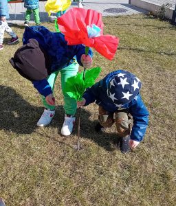 Dwóch chłopców na przedszkolnym trawniku "sadzi" czerwonego kwiatka zrobionego z bibuły.