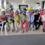 Dziewczynki siedzą na krzesłach i trzymają w ręku kwiatek zrobiony z balonów.