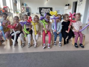 Dziewczynki siedzą na krzesłach i trzymają w ręku kwiatek zrobiony z balonów.