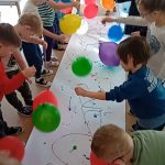 Dzieci stoją dookoła długiego arkusza białego papieru. W rączkach trzymają balony na sznurkach , którymi stemplują kolorową farbkę, rozmieszczoną na arkuszu.